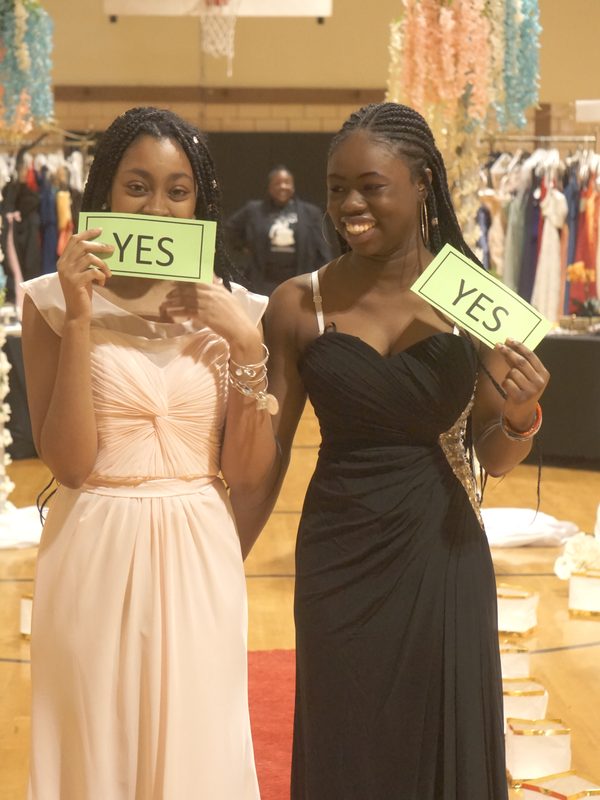 Two young women in prom gowns holding YES signs on the red carpet at the Prom Boutique